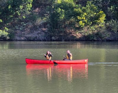 Two people in a bright red canoe paddling on a calm lake with a tree-lined shore in the background.
