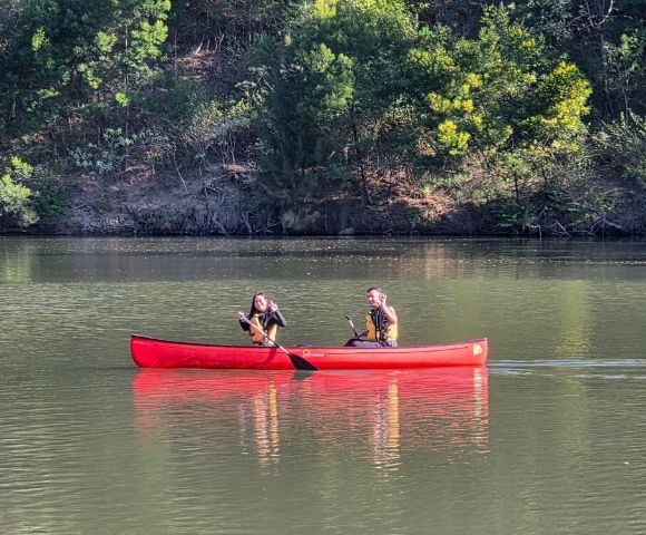 Two people in a bright red canoe paddling on a calm lake with a tree-lined shore in the background.