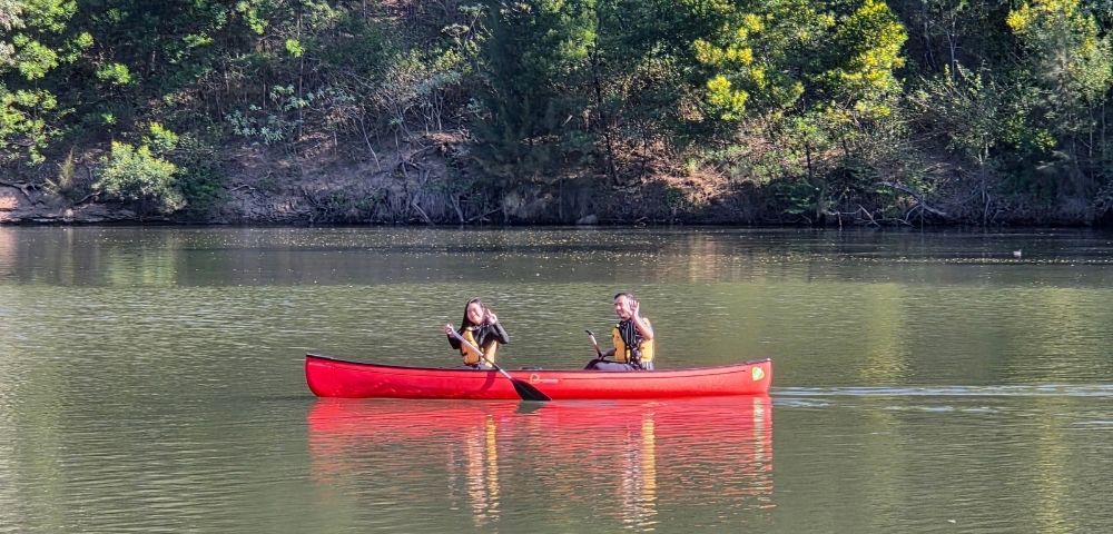 Two people in a bright red canoe paddling on a calm lake with a tree-lined shore in the background.