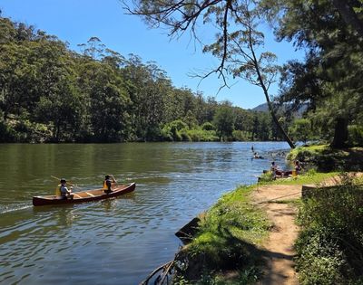 Two individuals paddle a canoe on a calm river with lush green trees covering the surrounding hillsides under a clear blue sky.