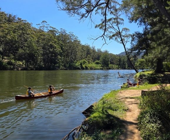 Two individuals paddle a canoe on a calm river with lush green trees covering the surrounding hillsides under a clear blue sky.