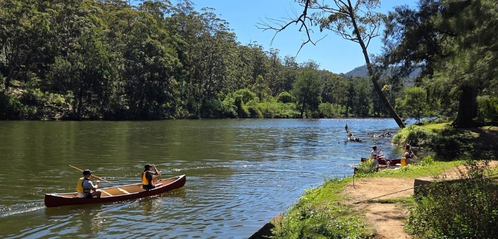 Two individuals paddle a canoe on a calm river with lush green trees covering the surrounding hillsides under a clear blue sky.