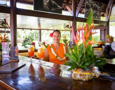 A smiling woman in an orange dress and flower lei stands behind a polished tropical bar, with colorful cocktails and greenery in the foreground.