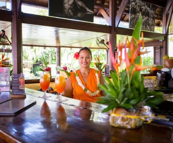 A smiling woman in an orange dress and flower lei stands behind a polished tropical bar, with colorful cocktails and greenery in the foreground.