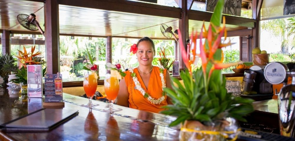 A smiling woman in an orange dress and flower lei stands behind a polished tropical bar, with colorful cocktails and greenery in the foreground.