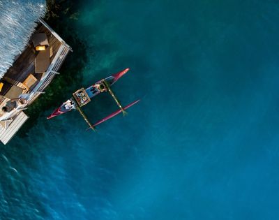 Aerial view of a small outrigger canoe beside a wooden boat on vivid turquoise water, evoking a calm, sunlit day at sea.