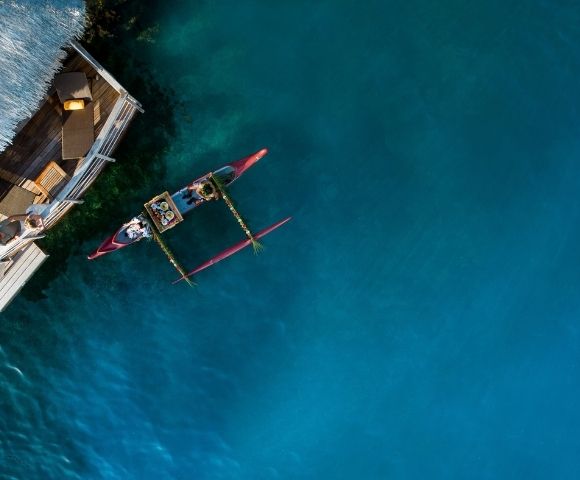 Aerial view of a small outrigger canoe beside a wooden boat on vivid turquoise water, evoking a calm, sunlit day at sea.