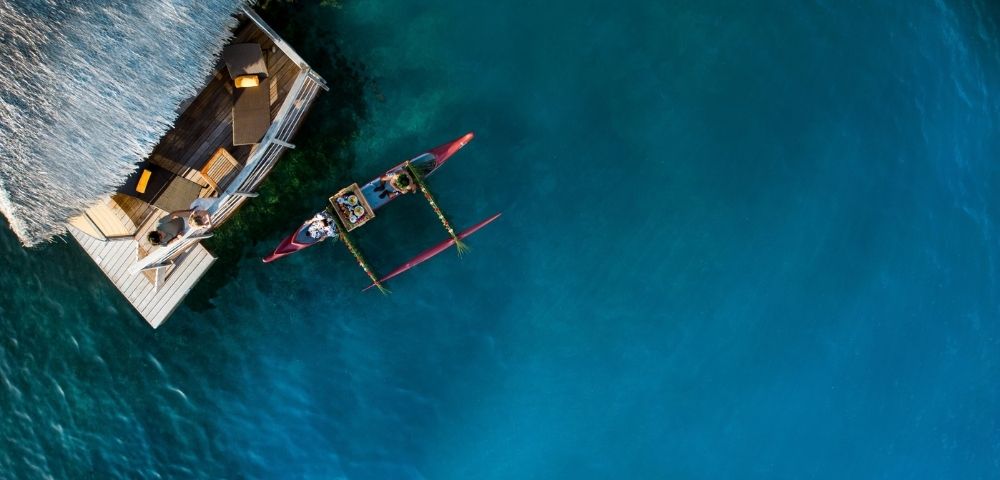 Aerial view of a small outrigger canoe beside a wooden boat on vivid turquoise water, evoking a calm, sunlit day at sea.