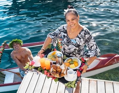 A smiling woman in a floral dress serves a colorful tropical meal from a narrow canoe, beside a cheerful man on sunlit blue water.