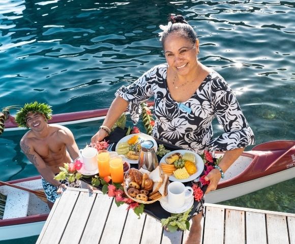 A smiling woman in a floral dress serves a colorful tropical meal from a narrow canoe, beside a cheerful man on sunlit blue water.