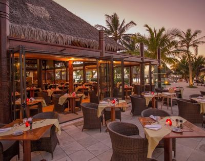 An open-air beachfront restaurant glows at dusk, with neatly set tables, wicker chairs, palm trees, and a calm, inviting tropical atmosphere.