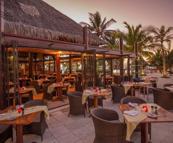 An open-air beachfront restaurant glows at dusk, with neatly set tables, wicker chairs, palm trees, and a calm, inviting tropical atmosphere.