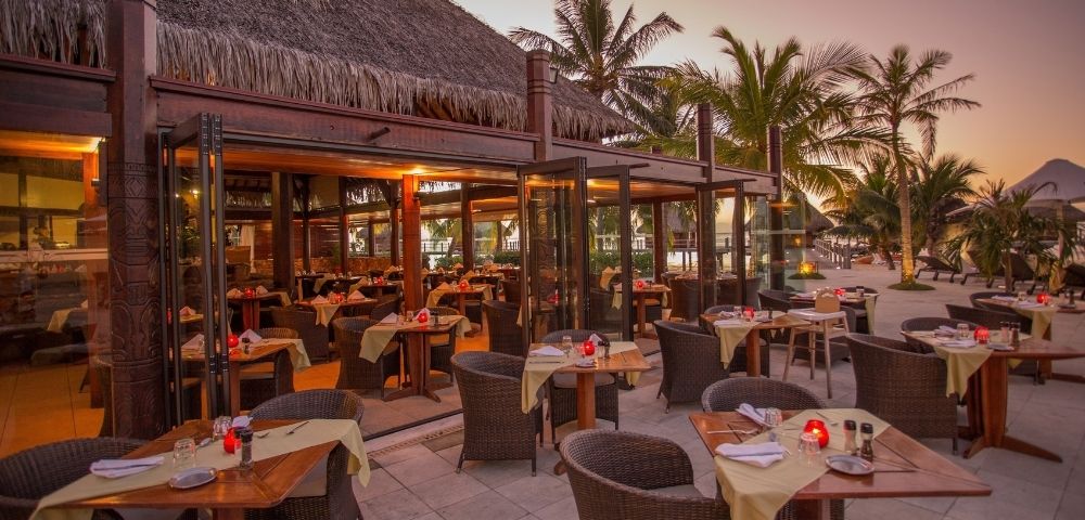 An open-air beachfront restaurant glows at dusk, with neatly set tables, wicker chairs, palm trees, and a calm, inviting tropical atmosphere.