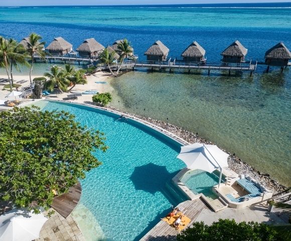 Aerial view of a tropical resort with a turquoise infinity pool, palm-lined beach, and overwater villas on clear blue water, creating a serene, luxurious mood.