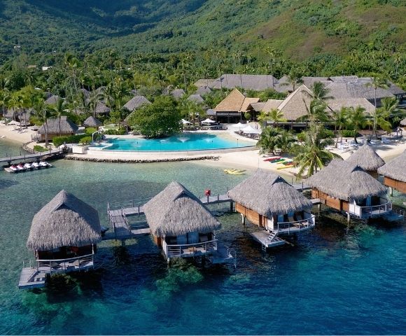 A tropical resort with thatched overwater bungalows above clear turquoise water, a sandy beach, palm trees, and green hills; serene and inviting.