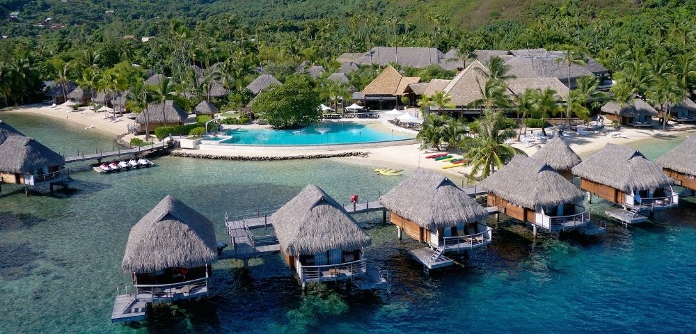 A tropical resort with thatched overwater bungalows above clear turquoise water, a sandy beach, palm trees, and green hills; serene and inviting.