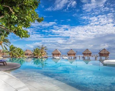 A tranquil tropical resort pool reflects thatched overwater bungalows beneath a bright blue, cloud-streaked sky, with palms and loungers framing the scene.