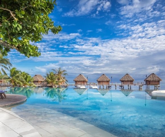 A tranquil tropical resort pool reflects thatched overwater bungalows beneath a bright blue, cloud-streaked sky, with palms and loungers framing the scene.