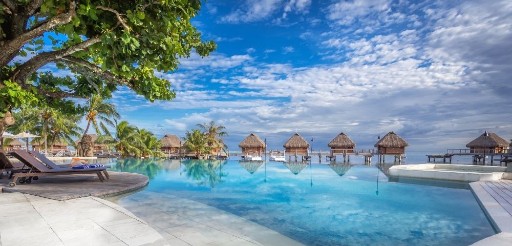 A tranquil tropical resort pool reflects thatched overwater bungalows beneath a bright blue, cloud-streaked sky, with palms and loungers framing the scene.