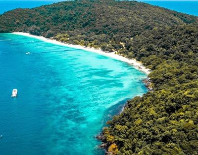 Aerial view of a tropical island coastline with a white sand beach, turquoise water, lush green forest, and a boat.