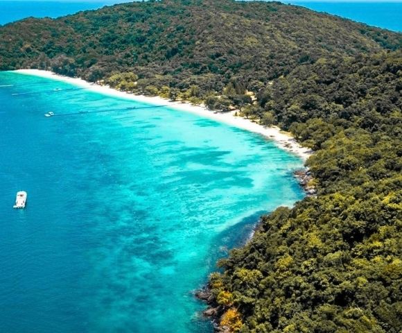 Aerial view of a tropical island coastline with a white sand beach, turquoise water, lush green forest, and a boat.