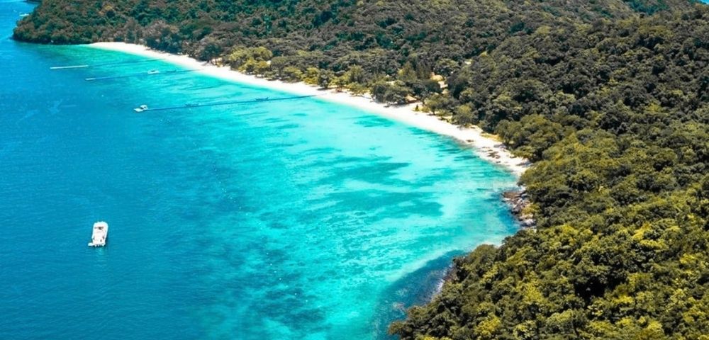 Aerial view of a tropical island coastline with a white sand beach, turquoise water, lush green forest, and a boat.