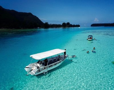 A white canopy boat floats on a brilliant turquoise lagoon, while swimmers and a smaller boat explore the clear water near lush island shores.