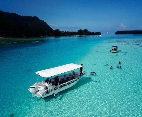 A white canopy boat floats on a brilliant turquoise lagoon, while swimmers and a smaller boat explore the clear water near lush island shores.