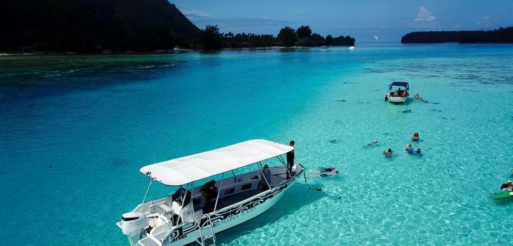 A white canopy boat floats on a brilliant turquoise lagoon, while swimmers and a smaller boat explore the clear water near lush island shores.