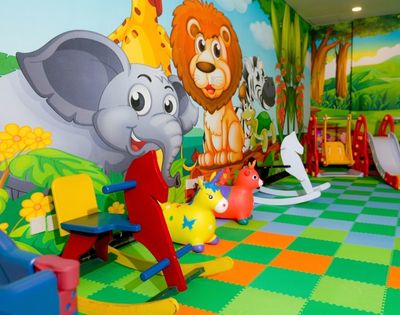 Colorful indoor jungle playroom with cartoon elephant, lion, and giraffe murals, animal toys, rocking seats, and slides on a bright checkerboard floor.