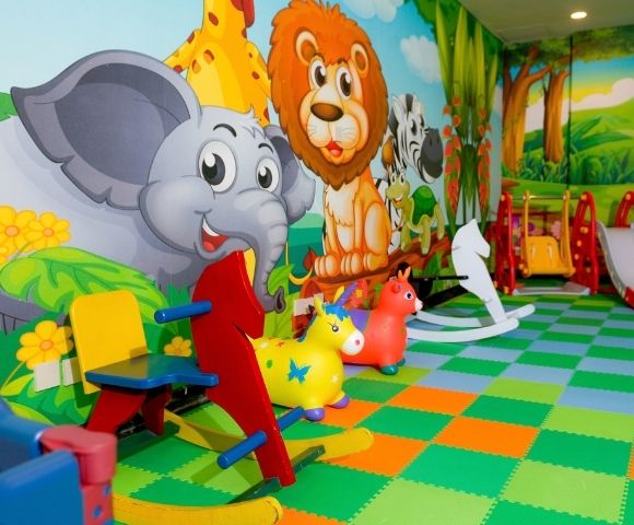 Colorful indoor jungle playroom with cartoon elephant, lion, and giraffe murals, animal toys, rocking seats, and slides on a bright checkerboard floor.