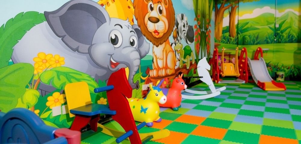Colorful indoor jungle playroom with cartoon elephant, lion, and giraffe murals, animal toys, rocking seats, and slides on a bright checkerboard floor.