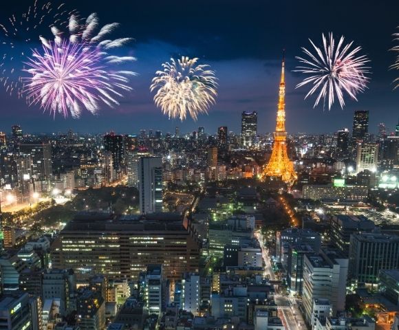 Nighttime aerial view of a sprawling cityscape illuminated by fireworks displays over the iconic Tokyo Tower.