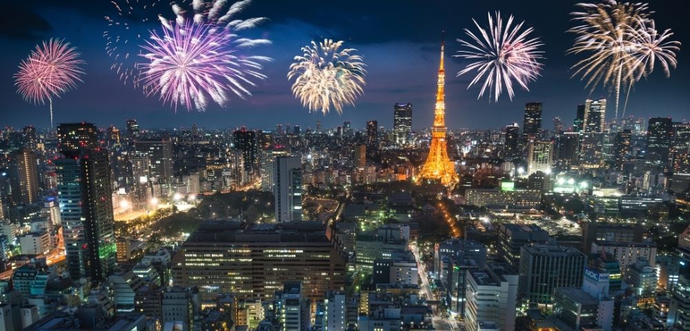 Nighttime aerial view of a sprawling cityscape illuminated by fireworks displays over the iconic Tokyo Tower.