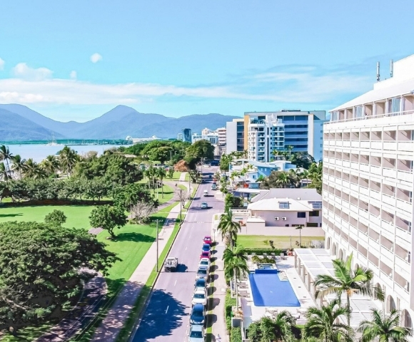 Aerial view of a coastal city street lined with palm trees, green parkland, and modern buildings, with a bay and mountains in the background under a bright blue sky.