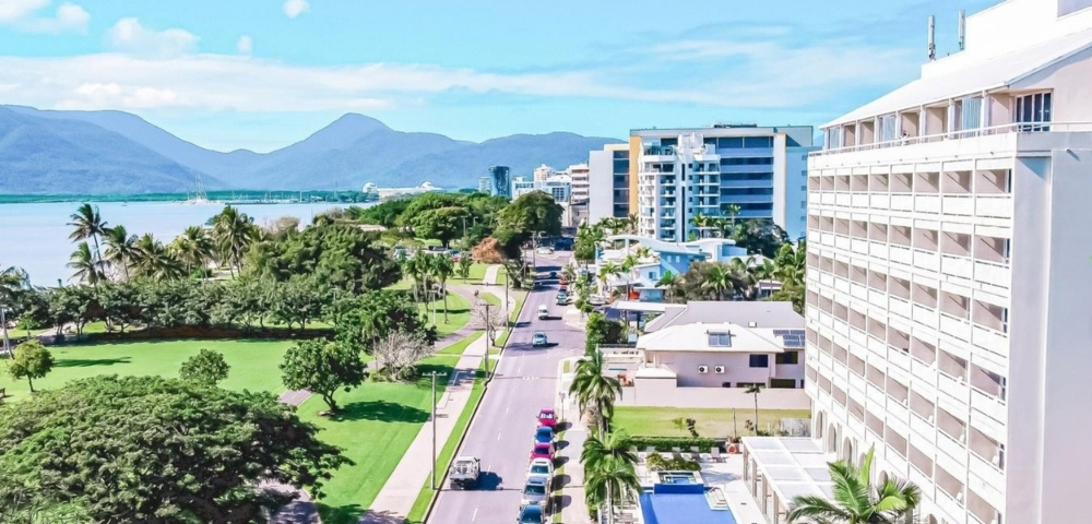 Aerial view of a coastal city street lined with palm trees, green parkland, and modern buildings, with a bay and mountains in the background under a bright blue sky.
