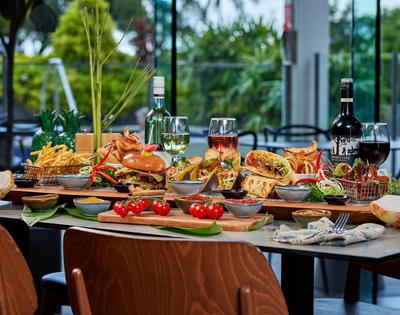 A bountiful platter of food featuring burgers, tacos, wraps, french fries, and various dips, with glasses of white and red wine on a table with a blurred outdoor background.