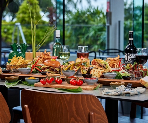 A bountiful platter of food featuring burgers, tacos, wraps, french fries, and various dips, with glasses of white and red wine on a table with a blurred outdoor background.