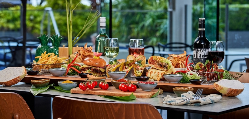A bountiful platter of food featuring burgers, tacos, wraps, french fries, and various dips, with glasses of white and red wine on a table with a blurred outdoor background.