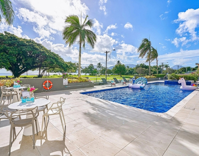 Outdoor swimming pool with inflatable unicorn and peacock floats, surrounded by lounge chairs and palm trees, with a view of the ocean and mountains in the background.
