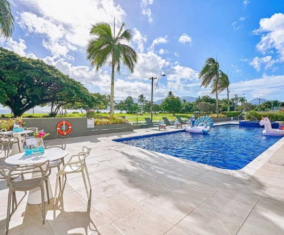 Outdoor swimming pool with inflatable unicorn and peacock floats, surrounded by lounge chairs and palm trees, with a view of the ocean and mountains in the background.
