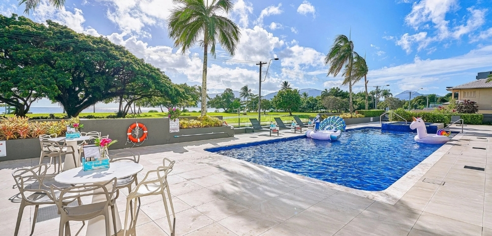 Outdoor swimming pool with inflatable unicorn and peacock floats, surrounded by lounge chairs and palm trees, with a view of the ocean and mountains in the background.
