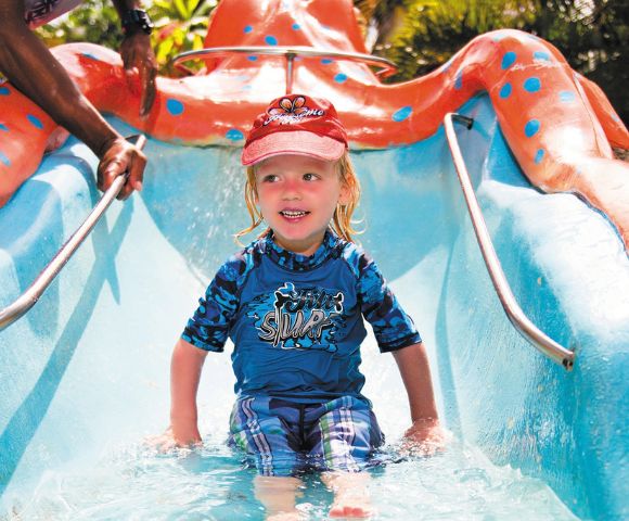 A young child sits at the bottom of a bright blue and orange water slide designed to look like an octopus, with hands reaching in from the sides of the frame.