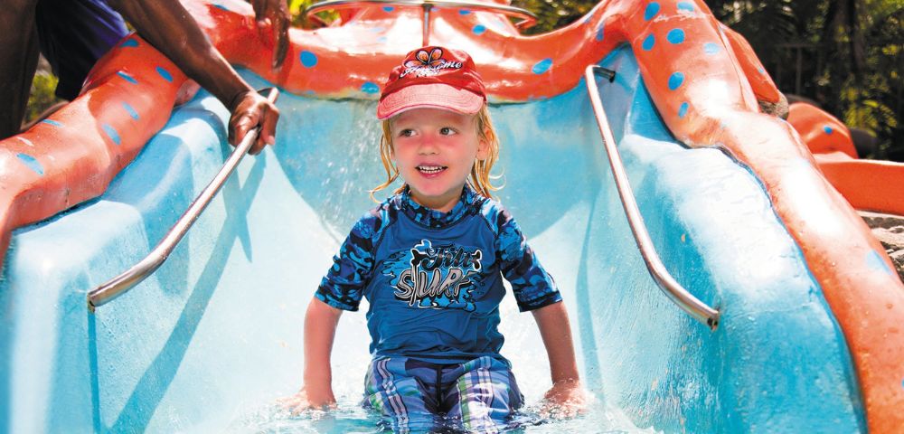A young child sits at the bottom of a bright blue and orange water slide designed to look like an octopus, with hands reaching in from the sides of the frame.