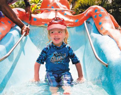 A young child sits at the bottom of a bright blue and orange water slide designed to look like an octopus, with hands reaching in from the sides of the frame.