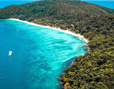 Aerial view of a lush tropical island, where dense green forest meets a curved white-sand beach and bright turquoise water; a small boat floats offshore.