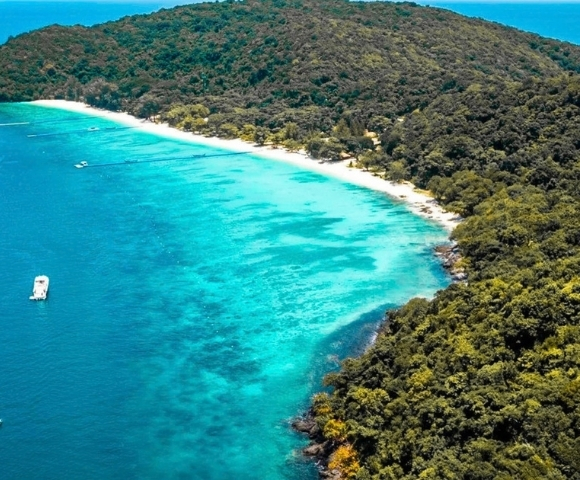Aerial view of a lush tropical island, where dense green forest meets a curved white-sand beach and bright turquoise water; a small boat floats offshore.