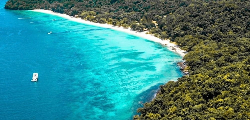 Aerial view of a lush tropical island, where dense green forest meets a curved white-sand beach and bright turquoise water; a small boat floats offshore.