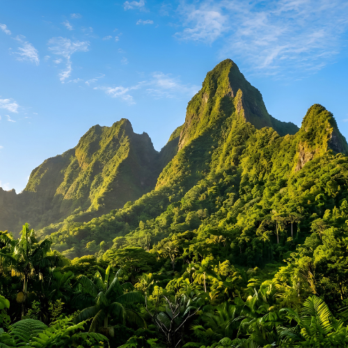 A dramatic series of steep, green mountain peaks rises above a dense tropical rainforest, illuminated by warm sunlight beneath a bright blue sky with scattered white clouds. Lush tropical vegetation fills the foreground, creating a vibrant landscape of rich greens and rugged volcanic-looking ridges.