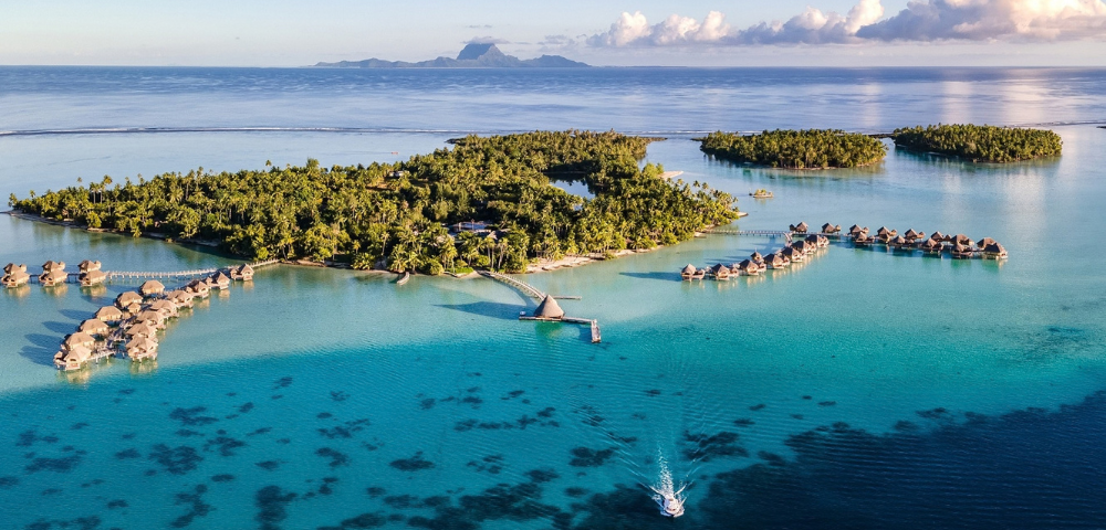 Aerial view of a lush tropical island surrounded by crystal-clear turquoise lagoon waters, with rows of overwater bungalows extending from the shoreline on curved wooden walkways. A small boat travels across the lagoon in the foreground, while distant mountainous islands and scattered clouds sit on the horizon under a bright blue sky.